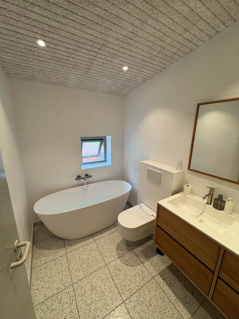 Lyst badeværelse med fritstående badekar, terrazzogulv, væghængt toilet og badmøbel i egetræ