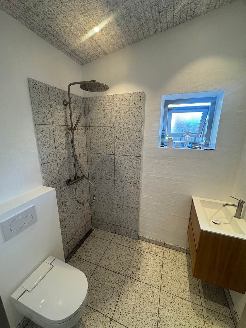 Badeværelse med åben bruseniche i terrazzofliser, regnbruser, væghængt toilet og håndvask i egetræ